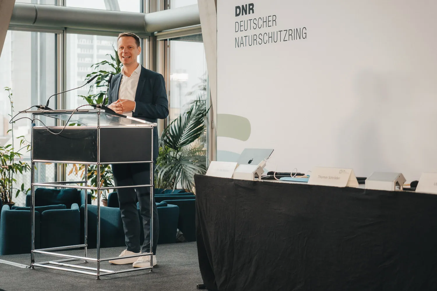 Blick auf das Podium und ein Stehpult, Kai Niebert begrüßt die Gäste, im Hintergrund eine Leinwand mit Logo und Aufschrift "DNR Deutscher Naturschutzring"